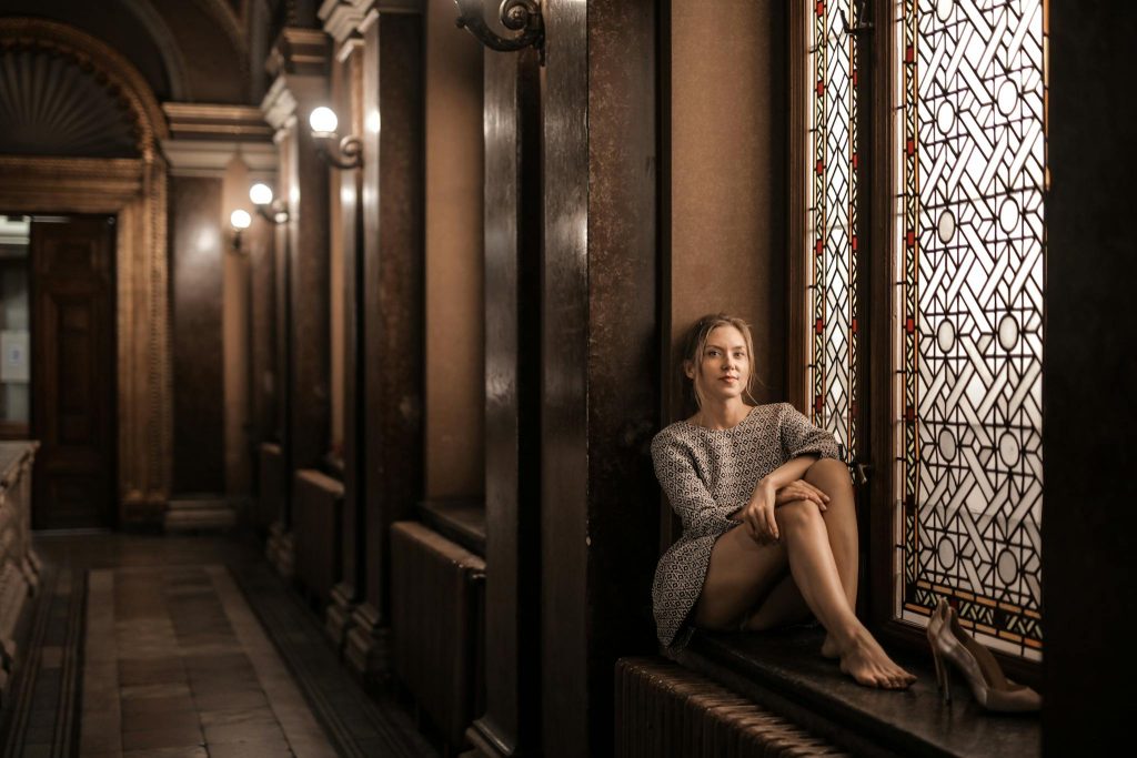 Blonde barefoot female sitting by painted window in dark lobby with Gothic interior