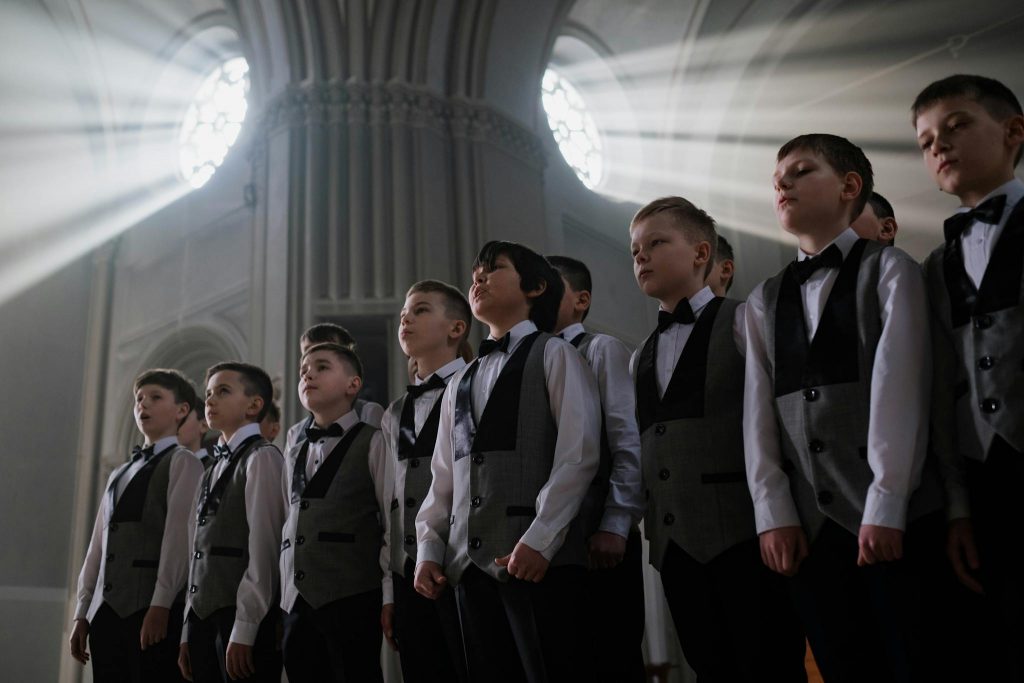 A group of boys in formal attire singing in a cathedral under dramatic sunlight.